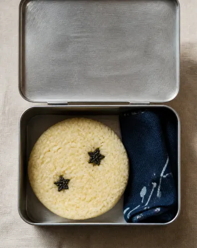 Natural-light analog still-life photography artwork: An aluminum lunchbox holds one round pale-yellow rice cake like a moon, two sesame stars and a folded navy cloth. The vertical composition preserves a clear subject and tactile handmade detail.