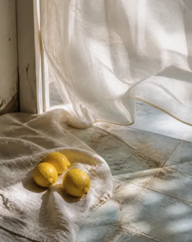Still-life and lifestyle photography / natural light artwork depicting three lemons beside rumpled linen as a sheer curtain moves over an old tiled floor. The vertical composition keeps the subject clear with tactile material and natural-light detail.